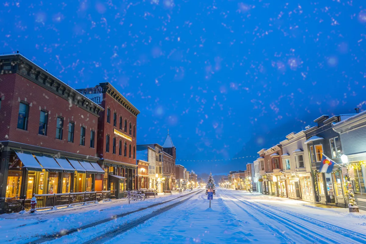A snow-covered street in Telluride lined with quaint, decorated shops beneath a twilight sky. A Christmas tree glows in the distance, and people stroll along the sidewalks, enjoying the festive atmosphere. The scene evokes a lively small-town nightlife and easy walkability, perfectly capturing the spirit of après-ski exploration.