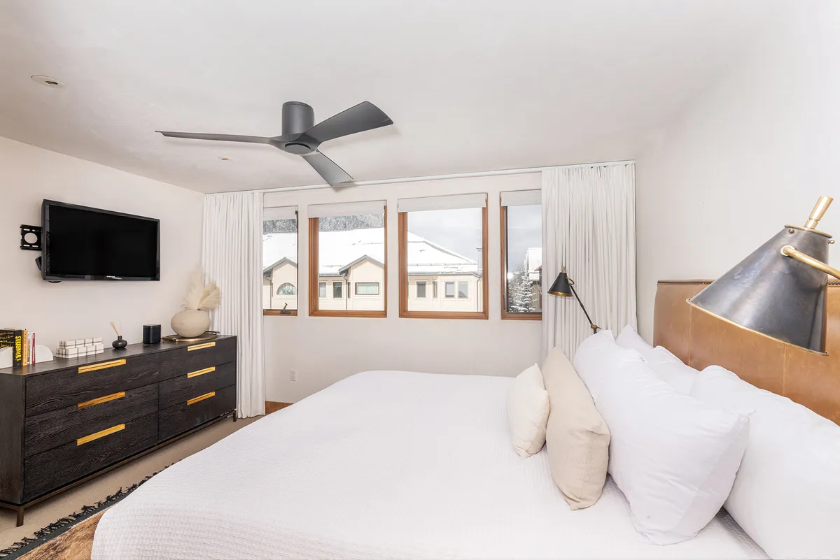 This image displays a bed with crisp white linens beside large windows that reveal stunning mountain views. A dark wood dresser, modern ceiling fan, and a brass-accented lamp add both style and comfort, while soft natural light enhances the room's tranquil, inviting mood.