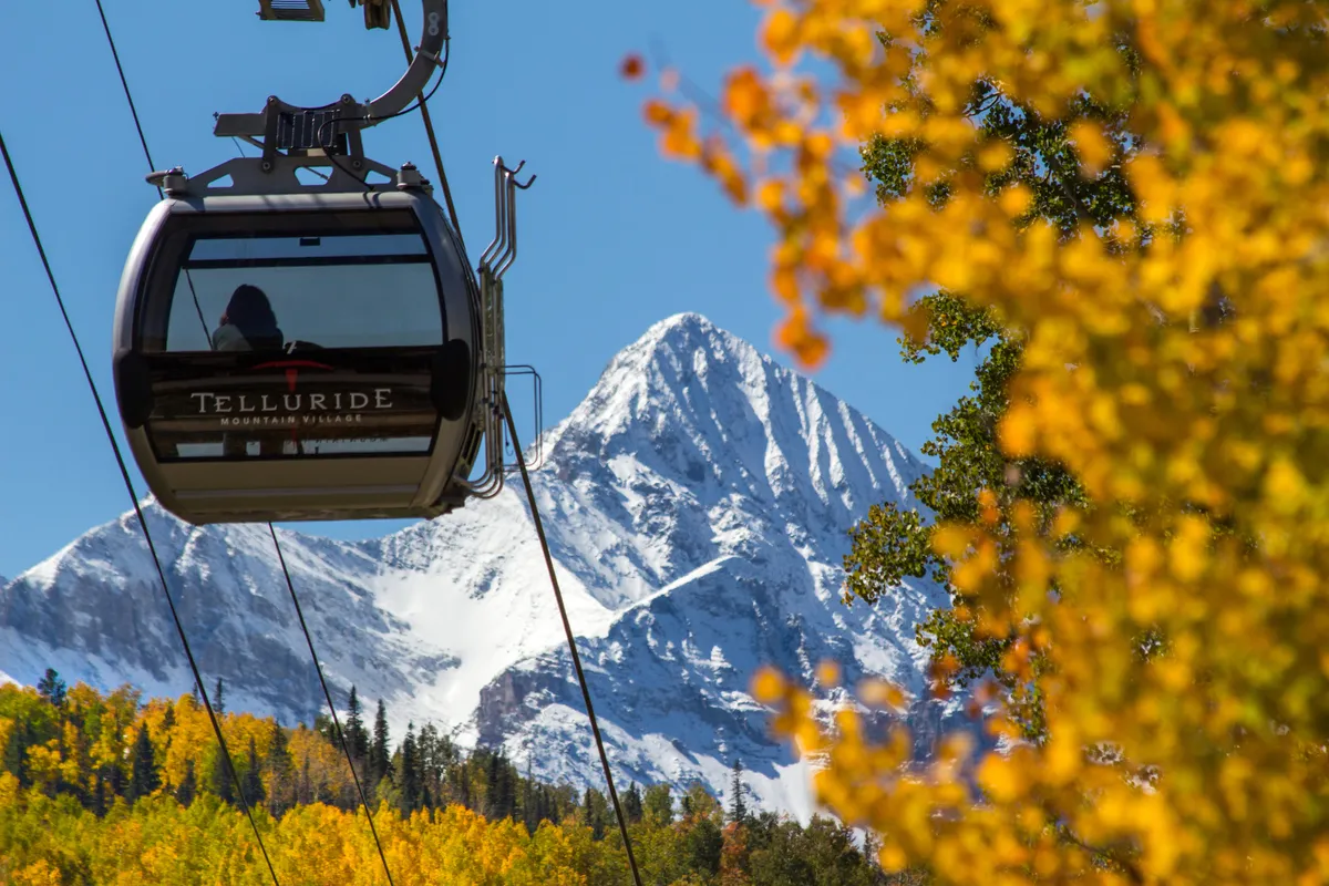 A sweeping exterior view featuring the iconic Telluride gondola gliding over colorful autumn foliage, with snow-capped mountains in the background. The image captures both the thrill of the ski season and the beauty of year-round outdoor adventures.