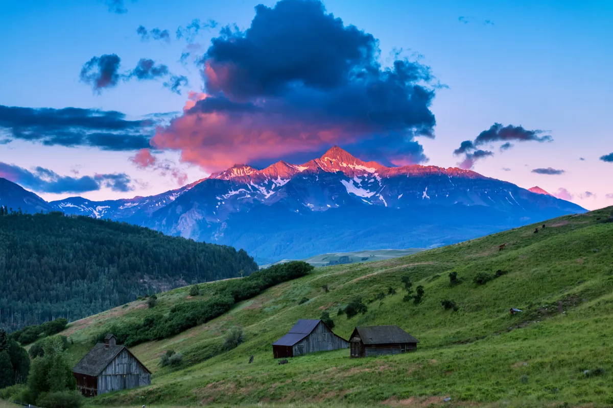 A breathtaking view of snow-capped mountains under a vibrant sunset sky, with rustic wooden cabins nestled in the landscape. The scene is bathed in warm light, suggesting a cozy yet adventurous mountain setting perfect for romantic escapes.