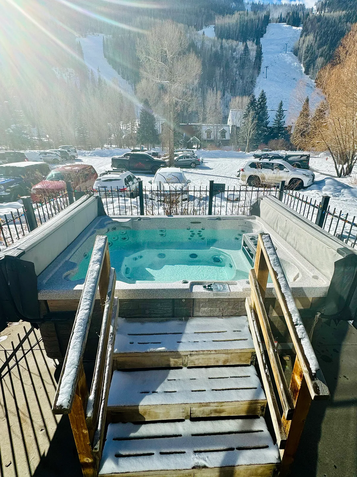 An inviting outdoor hot tub sits at the edge of snow-covered stairs, framed by stunning mountain views. Steam rises as friends gather in the warmth, contrasting with the crisp winter air and sunset skies.