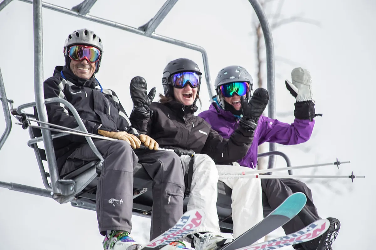 Three friends ascend a ski lift with snow-covered mountains in the background, each carrying ski equipment and wearing bright winter gear. The expressions and setting exude excitement, camaraderie, and anticipation for both adventure and après-ski socializing.