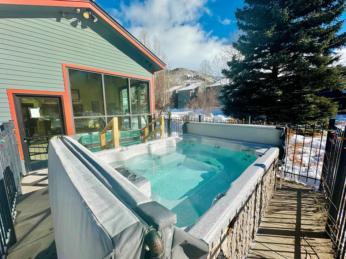 A modern exterior shot showing an outdoor hot tub, surrounded by lush greenery and set against a backdrop of mountains. The scene evokes a sense of tranquility, with the tub as a social focal point for relaxation or outdoor gatherings.