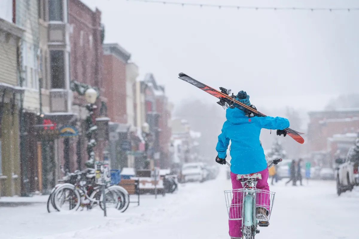 A charming winter street scene featuring colorful buildings, a person riding a bicycle with skis attached, and a fresh dusting of snow. The vibrant town atmosphere is lively but cozy, hinting at boutique shopping, cafes, and easy access to local amenities.