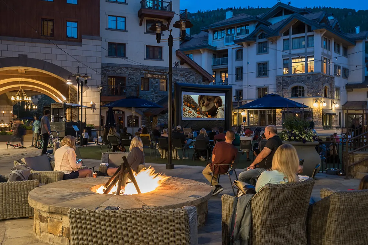 An inviting outdoor scene with a fire pit, an outdoor movie screen, and a cozy seating area set against a mountain backdrop. The setup is perfect for summer festivals, socializing, and enjoying cool evenings beneath the stars.