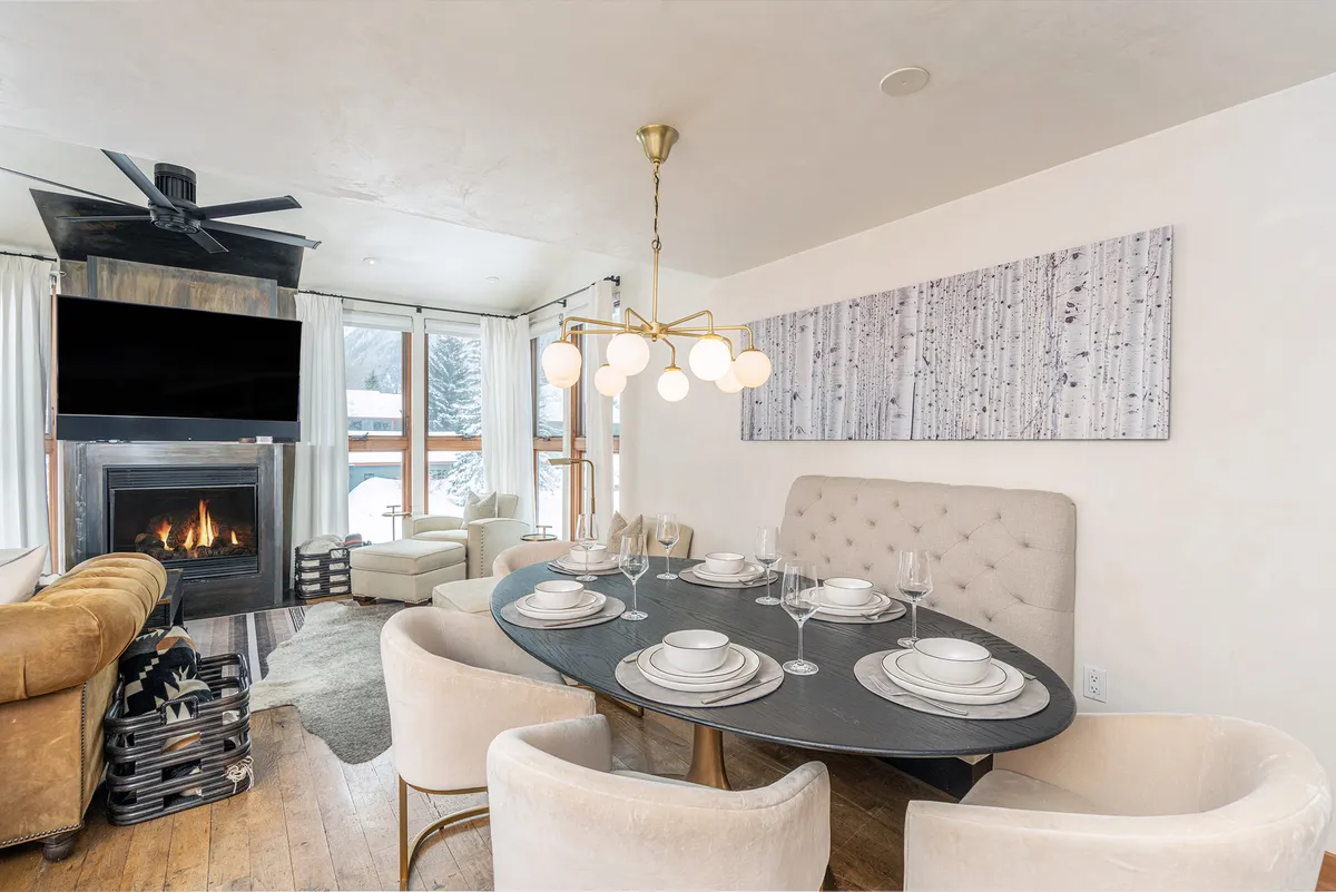 The image showcases a spacious, inviting living and dining area flooded with natural light from floor-to-ceiling windows. A large dining table is beautifully set for a meal, with comfortable seating nearby, a glowing fireplace, tasteful artwork, and mountain views visible through the windows. The scene projects warmth and togetherness.