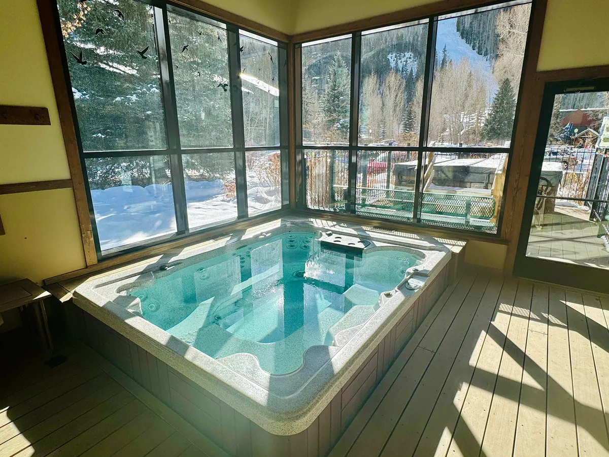 This image features a spacious indoor hot tub area with wooden flooring and large windows that offer mountain views. The space is designed for relaxation, with the inviting hot tub as the focal point, and the snowy landscape outside enhancing the cozy, resort-like ambiance.