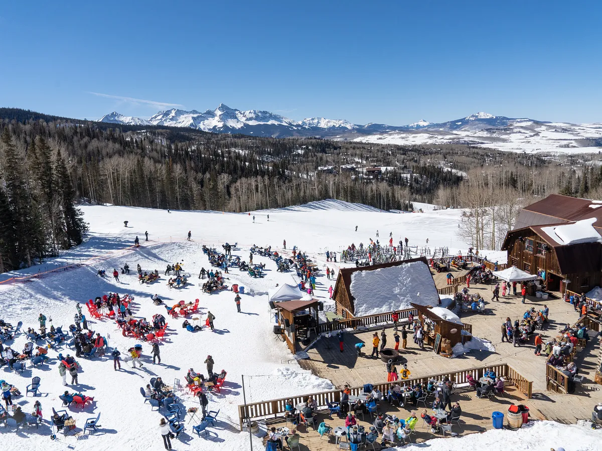 A lively scene at the base of the ski slopes features people skiing and relaxing on sunlit chairs, with the ski lodge and snow-capped mountains in the background. The energy and activity capture the excitement of a ski-in, ski-out experience, while the sunset glow adds warmth and a sense of adventure.