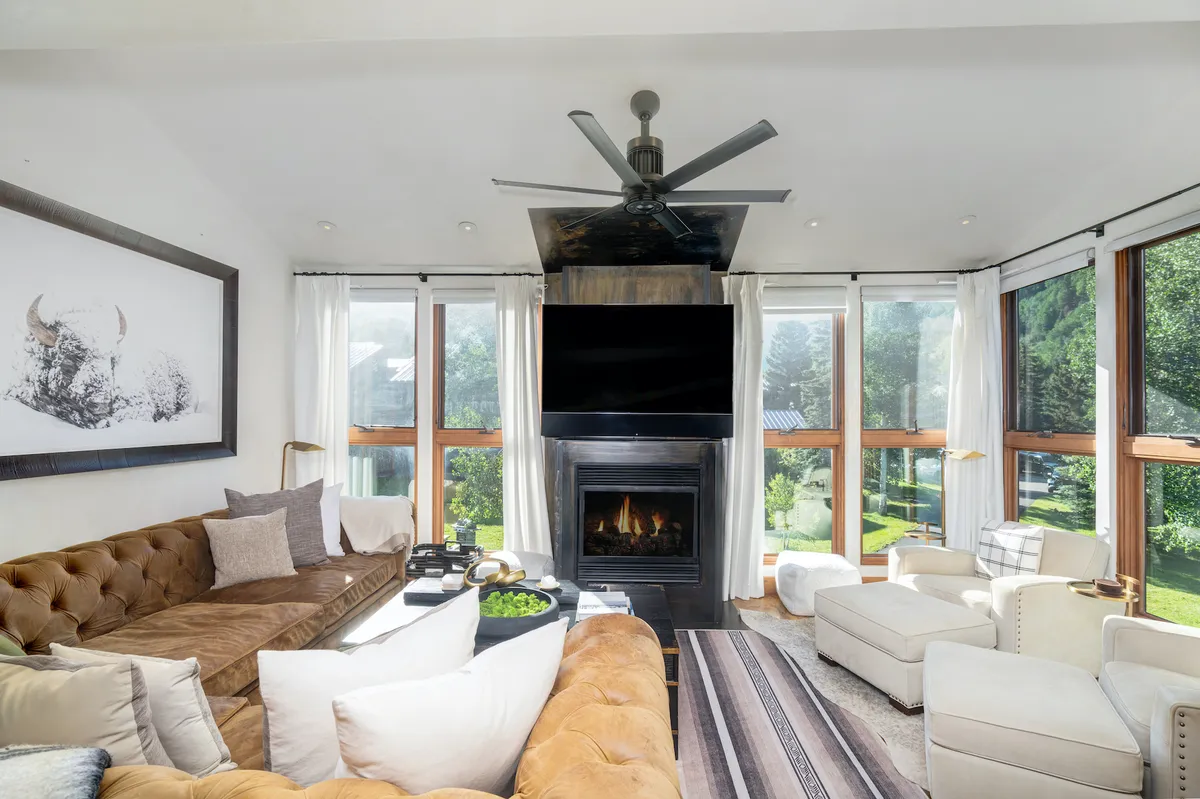 A spacious living room with a large sectional sofa oriented toward a modern fireplace. Expansive windows bring in natural light and frame partial views of the outdoors, while additional seating and artwork foster a cozy, welcoming vibe. The scene conveys relaxation and casual socializing.