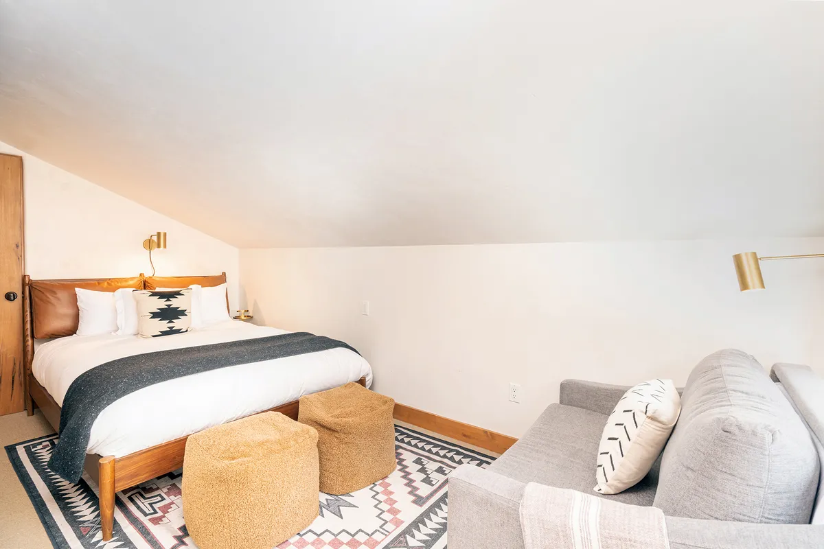 A bedroom with decorative pillows on a large bed, a golden bedside lamp, textured area rug, and a soft grey sofa with poufs nearby. Morning light streams in, creating an atmosphere perfect for relaxing, reading, or enjoying a quiet moment.