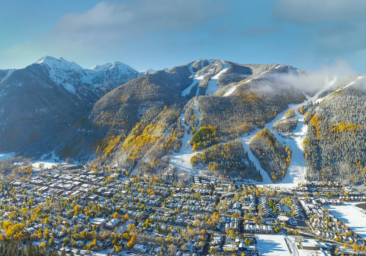 A panoramic view over Telluride showing snow-capped mountains, ski slopes, vibrant autumn trees, and the picturesque town below. The image seamlessly blends the thrill of winter sports with the charm and tranquility of off-season months.