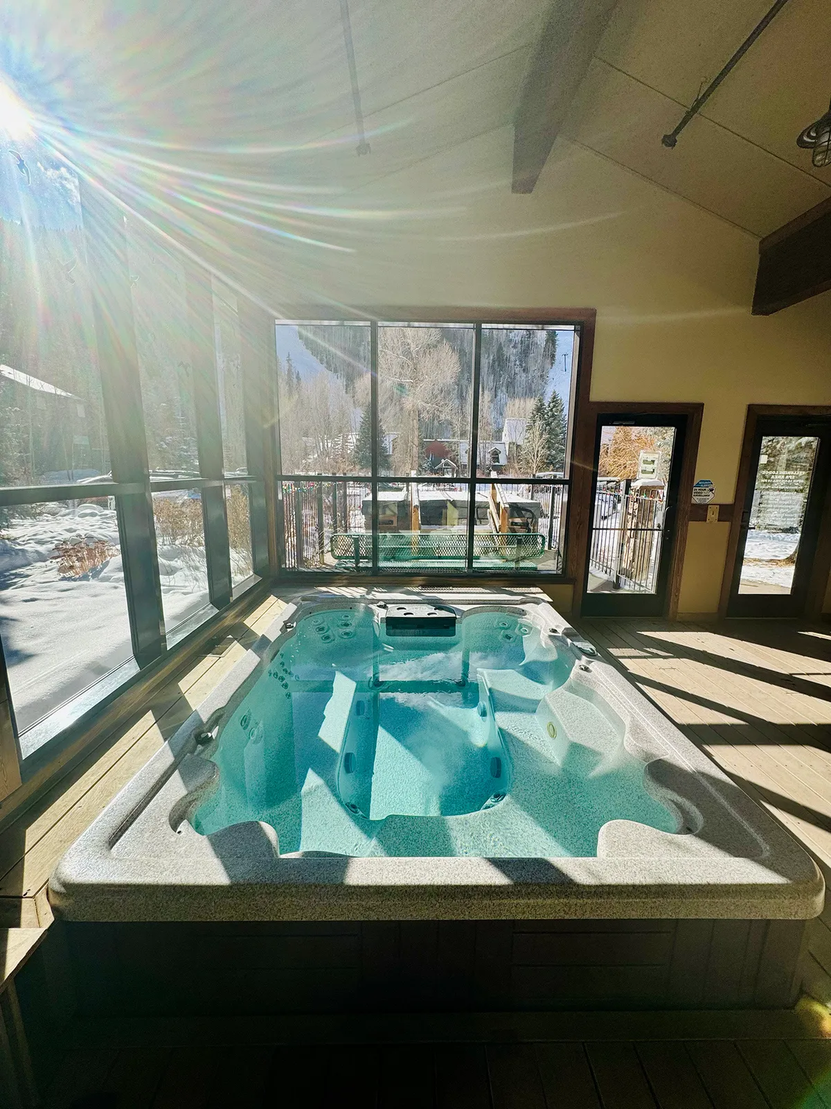 A tranquil indoor communal hot tub surrounded by large windows with a view of snow-dusted mountains. Warm lighting and comfortable seating provide an inviting space for soaking and relaxation against a dramatic winter backdrop. The setting is ideal for unwinding with friends after adventure-filled days.