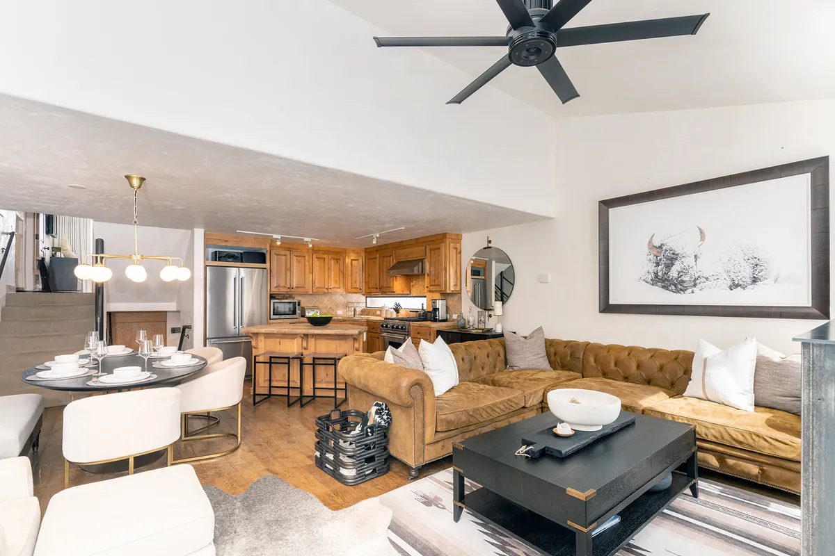 An inviting open-concept space featuring a chic sectional sofa, a modern dining table set, and a kitchen area seamlessly connected in the background. Large windows allow sunlight to pour in, brightening the entire interior and highlighting the comfortable and social layout. Tasteful artwork and decor complement the room’s warm, welcoming atmosphere.