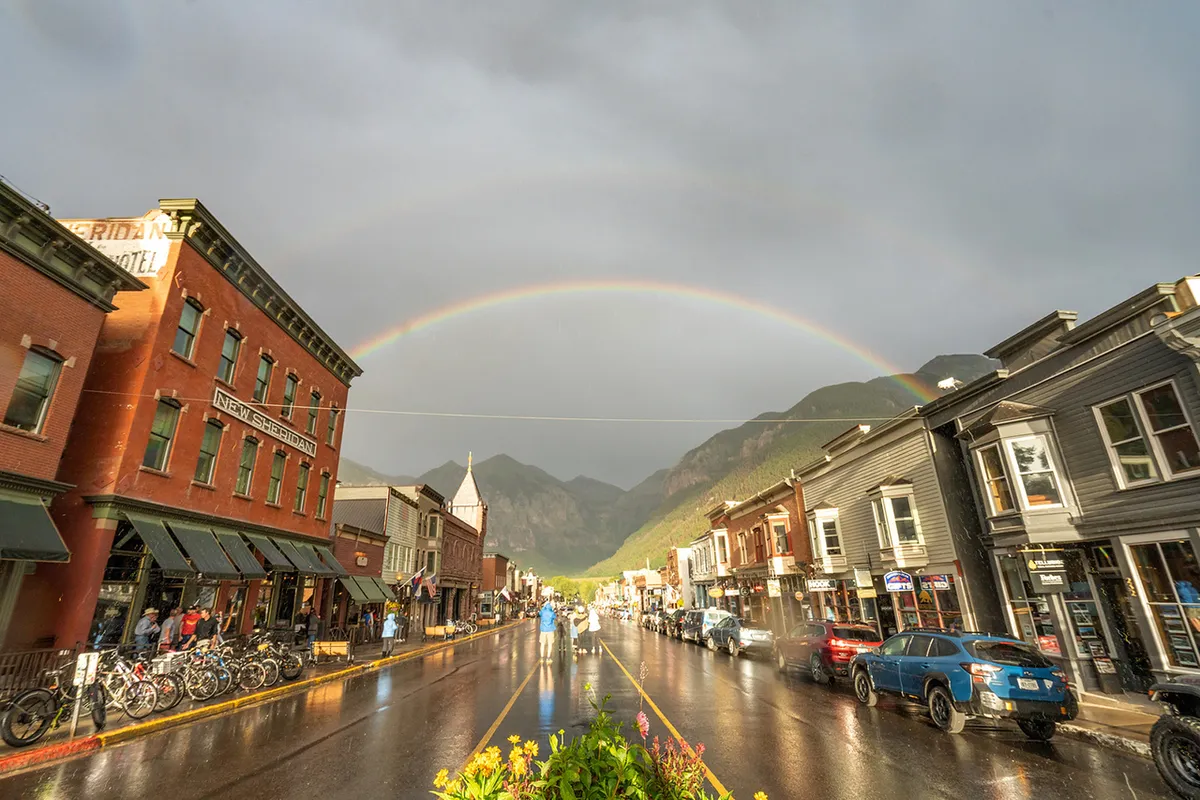 A post-rain scene in Telluride’s historic downtown: a rainbow arches over vibrant buildings and a glistening street. People stroll, shop, and enjoy the lively, welcoming town atmosphere with a mountain backdrop.